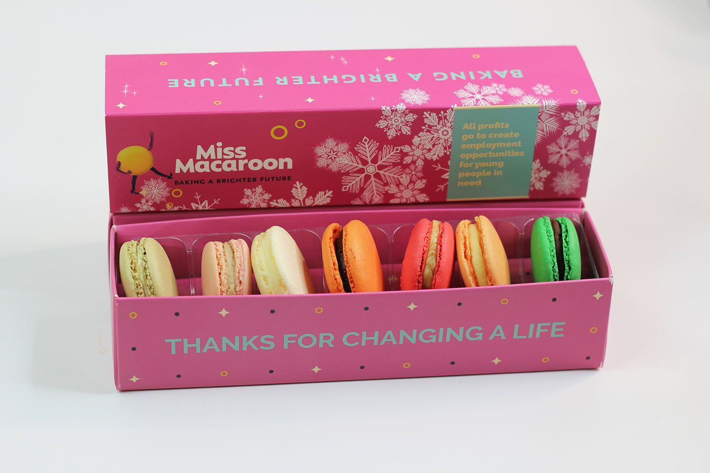 A bright pink gift box containing seven macaroons in bright colours. On the side of the gift box there is a message in blue 'Thanks for changing a life'. Behind the gift box there is a bright pink lid with the Miss Macaroon logo and a message that says 'All profits go to create employment opportunities for young people in need'.