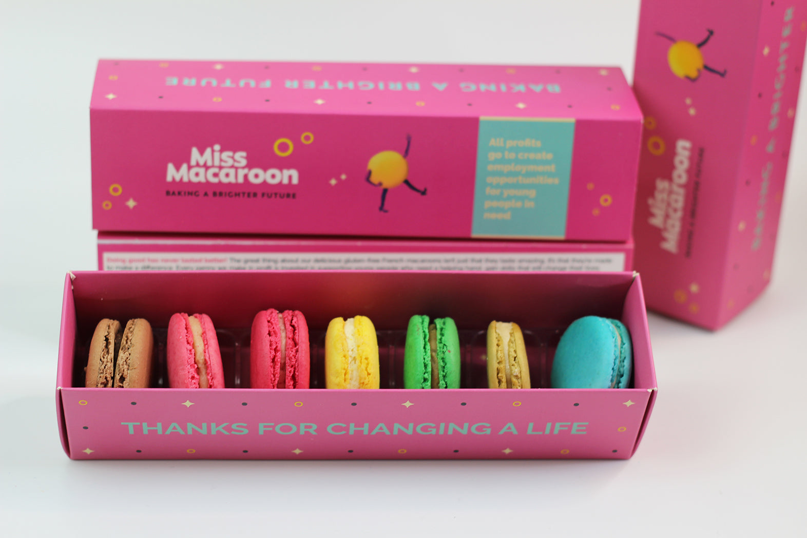 A bright pink gift box containing seven macaroons in bright colours. On the side of the gift box there is a message in blue 'Thanks for changing a life'. Behind the gift box there is a bright pink lid with the Miss Macaroon logo and a message that says 'All profits go to create employment opportunities for young people in need'.