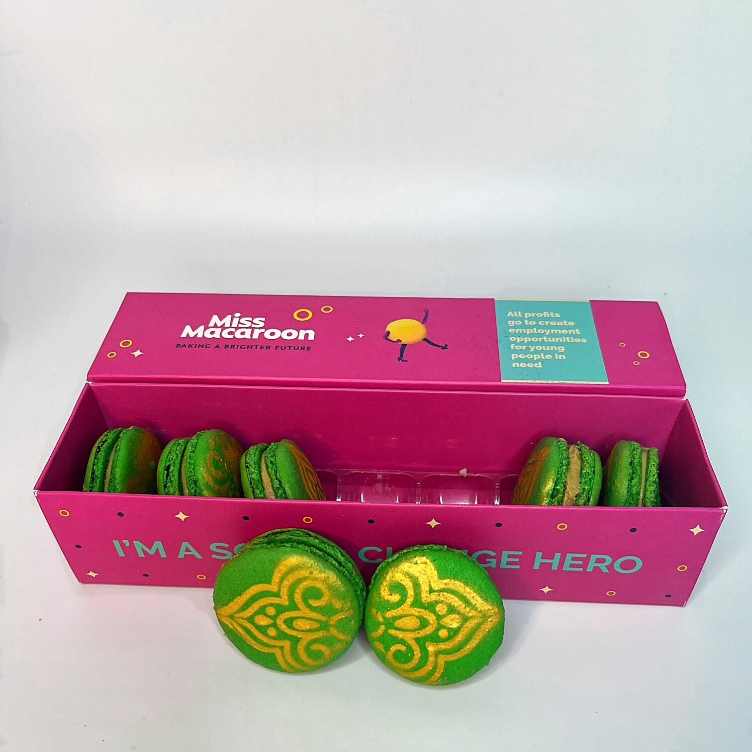 Bright pink gift box of seven green pistachio macaroons with an ornate gold pattern sprayed onto the macaroons. The gift box say 'I's a social change hero' in blue text on the side.